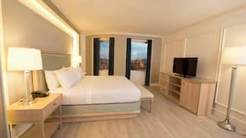 Ein Hotelzimmer mit Bett, Nachttischlampen, einem Fernseher und Blick auf die Stadt durch große Fenster.
