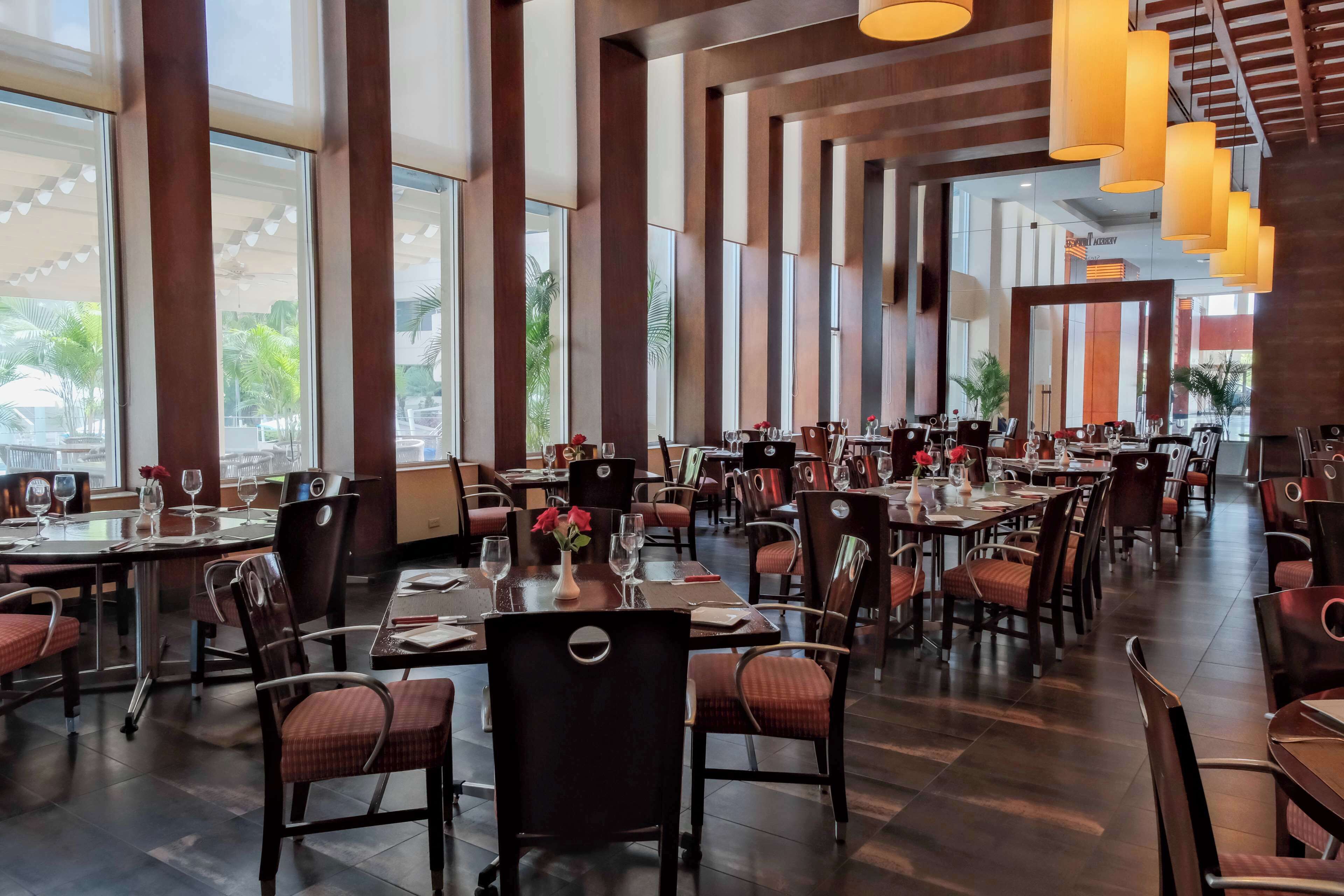 En rummelig restaurant med højt til loftet, store vinduer og moderne lysarmaturer. Bordene står pænt opstillet med stole, og indretningen har en blanding af mørkt og lyst træ.
