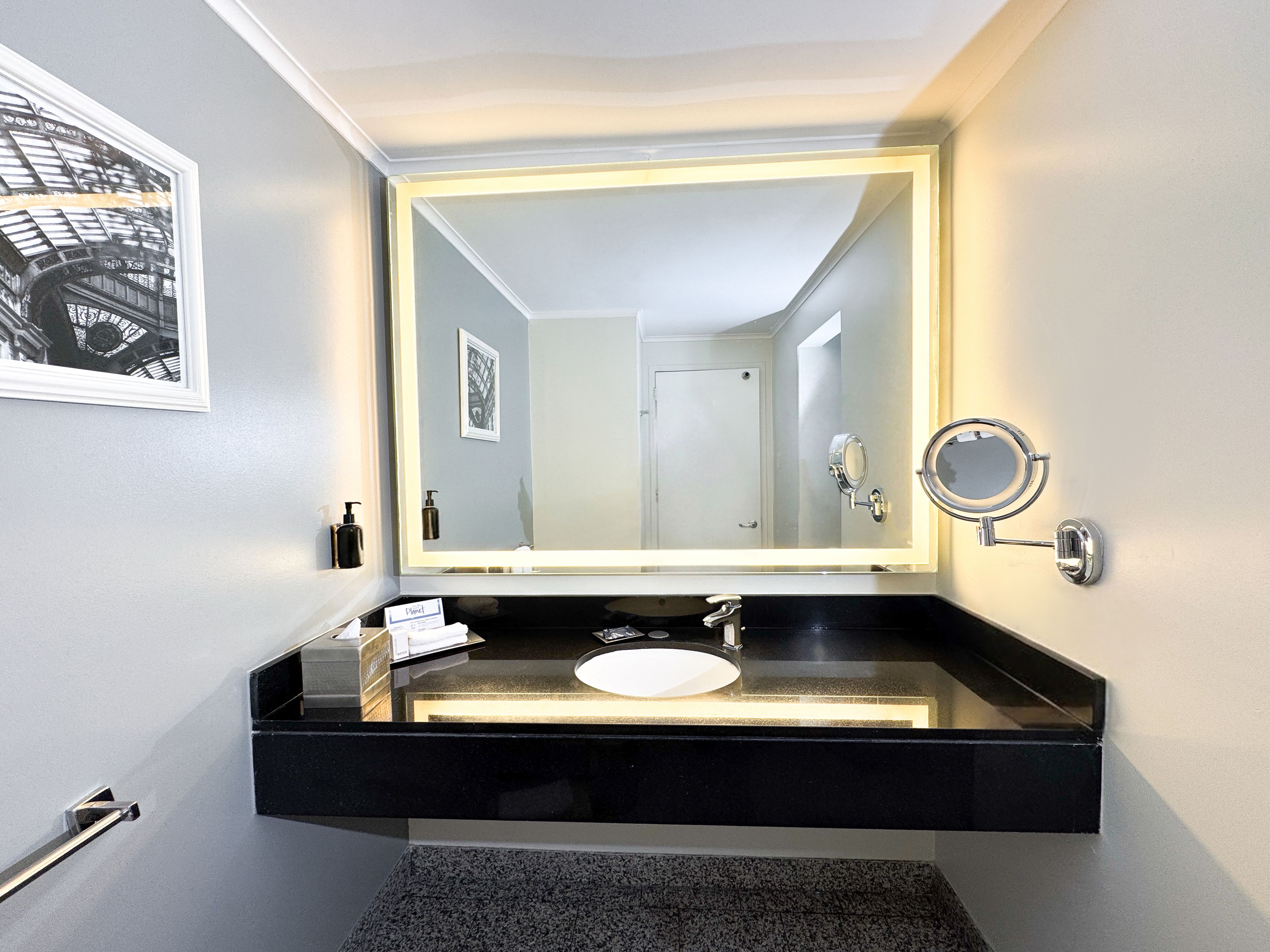 Un baño con un espejo grande, un lavamanos, un perchero para toallas y un cuadro en la pared.