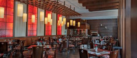 Un restaurante espacioso con varias mesas y sillas, techo de madera con luces colgantes y ventanas grandes con iluminación roja.