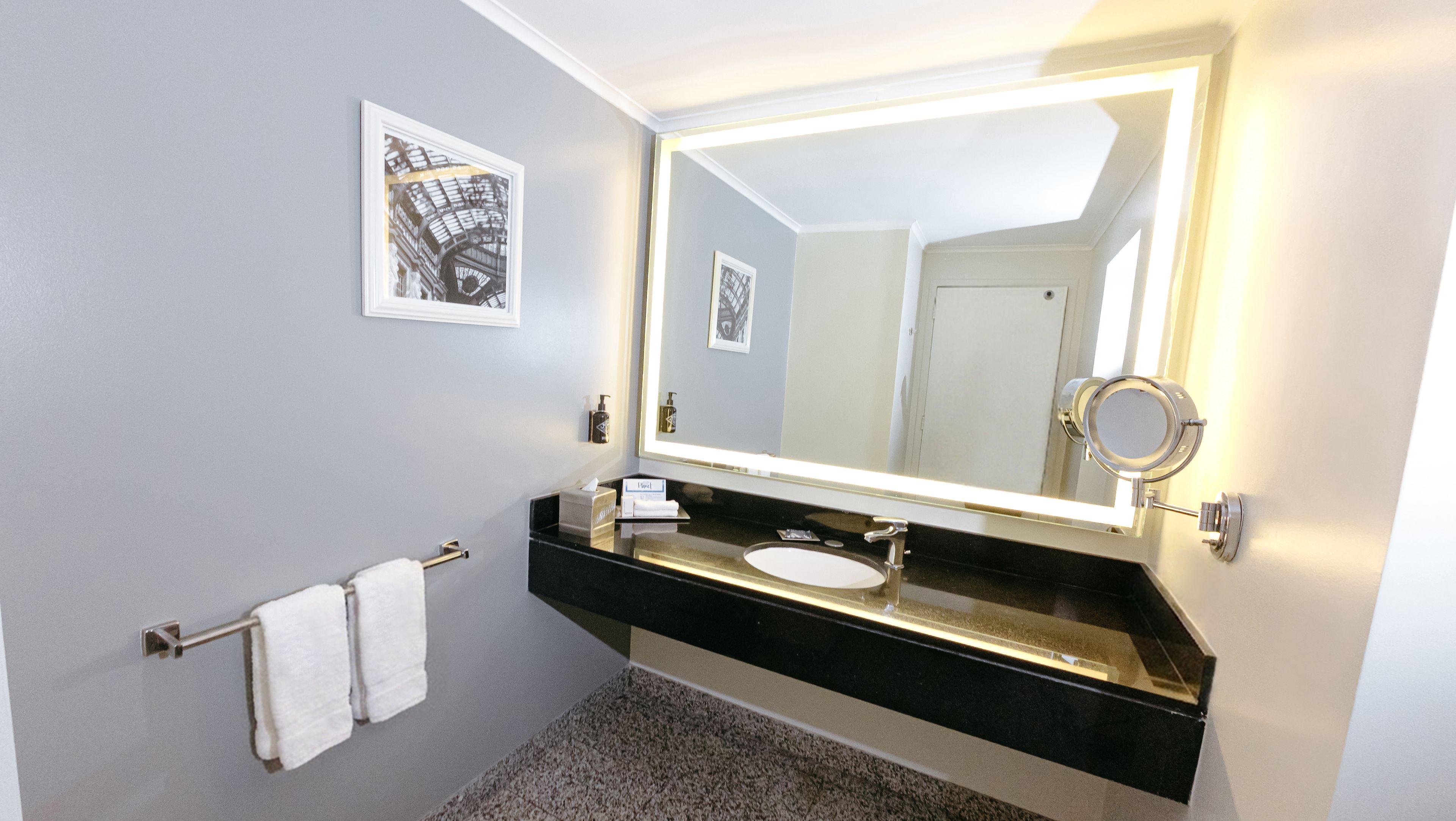 Un baño con encimera negra, lavamanos blanco, espejo grande con iluminación y dos cuadros en la pared.