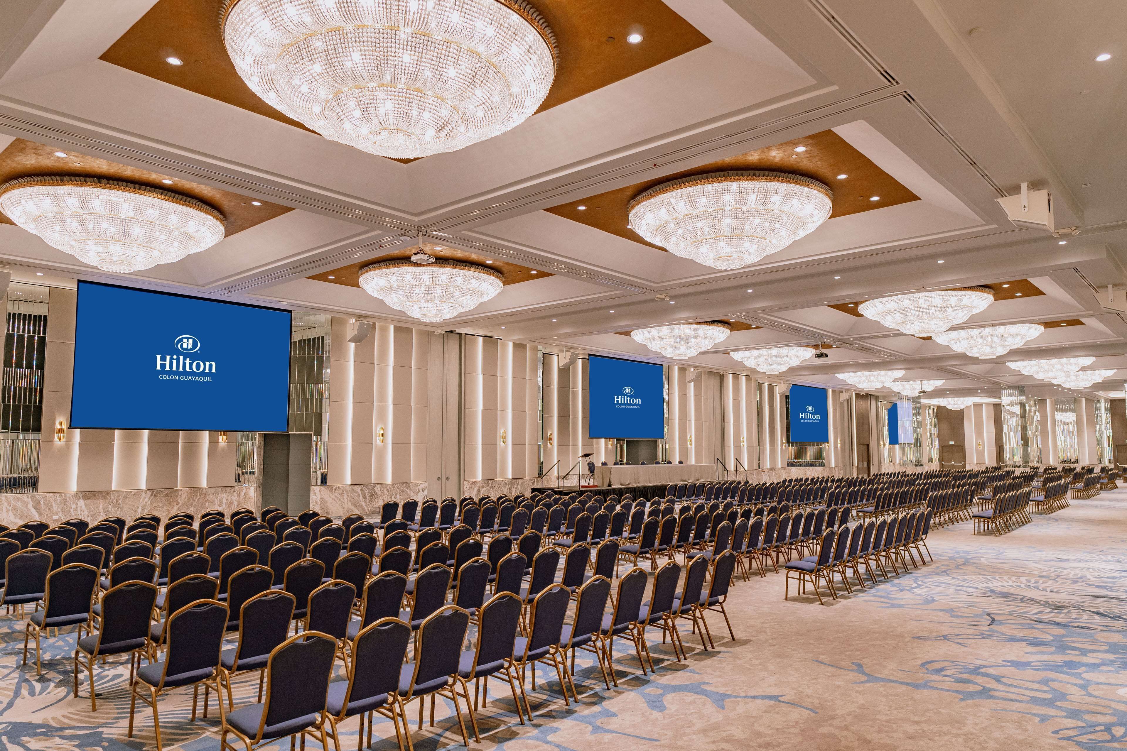 A large conference room with rows of chairs, a stage, and Hilton branding.