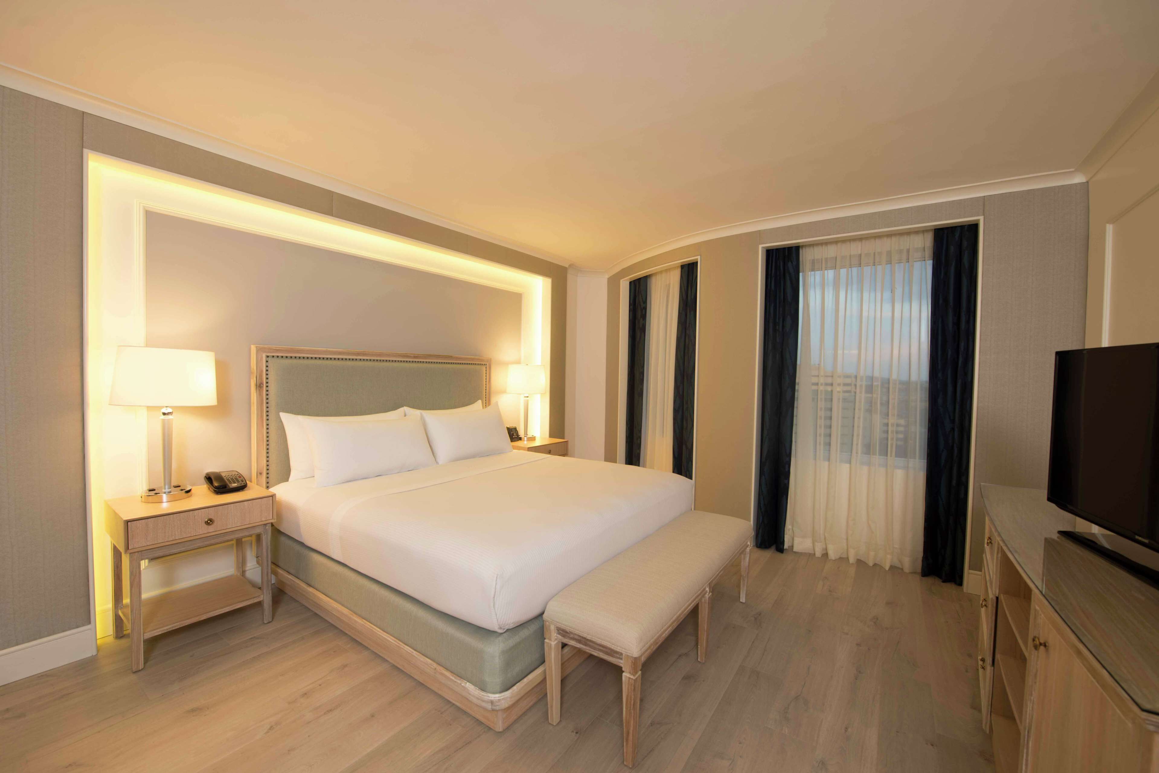 A hotel room with a large bed, bedside tables, a bench, a television, and a window with curtains.