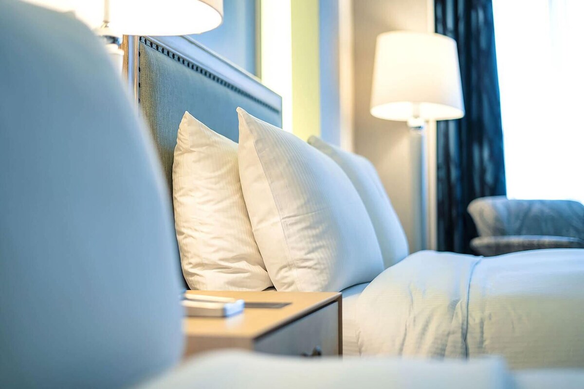 Ein ordentlich bezogenes Bett mit weißen Leinen und Kissen, ein Nachttisch mit einer Lampe und Blick durch ein mit Vorhängen drapiertes Fenster.