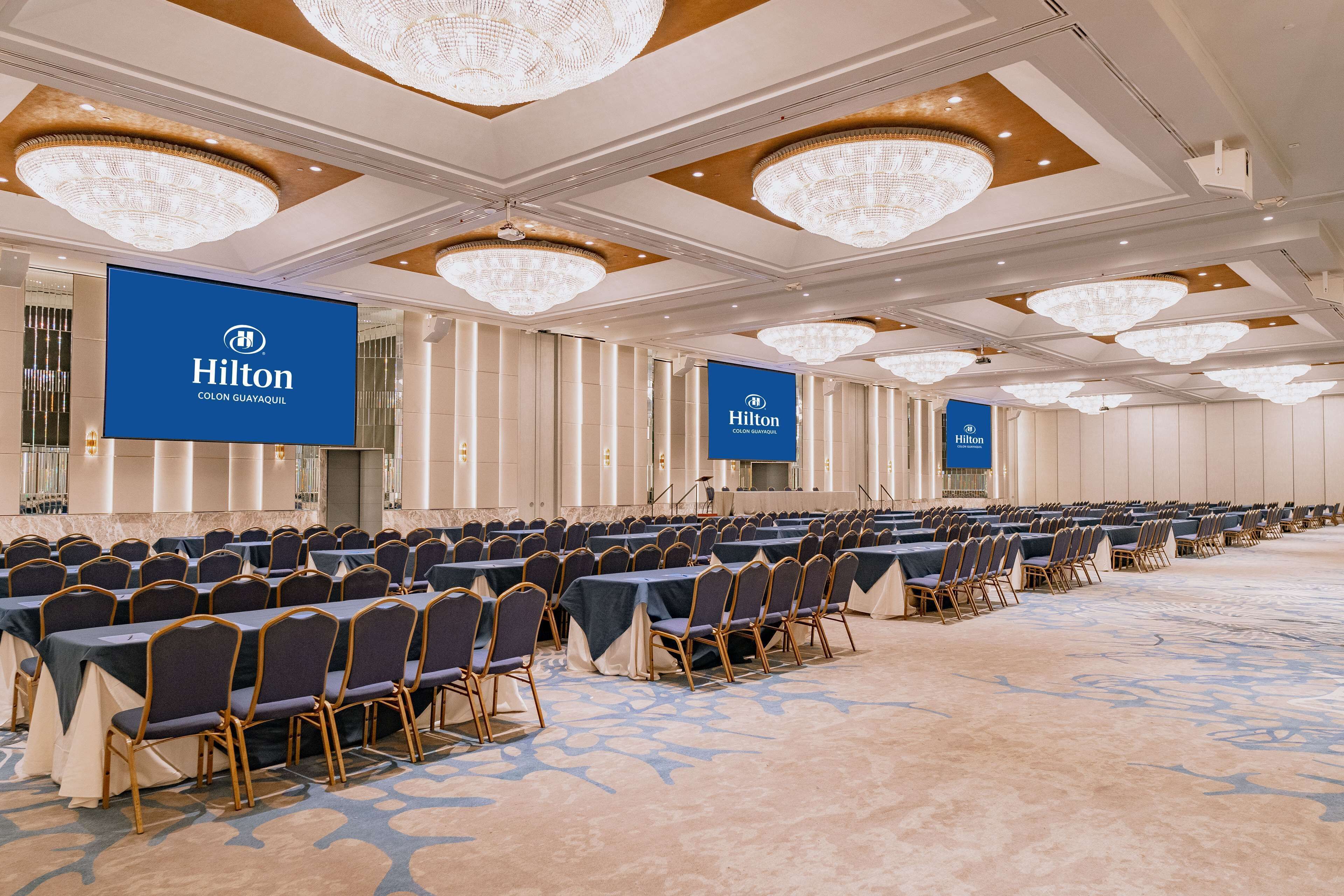 A spacious conference room with rows of tables and chairs, large chandeliers, and Hilton branding on the walls.