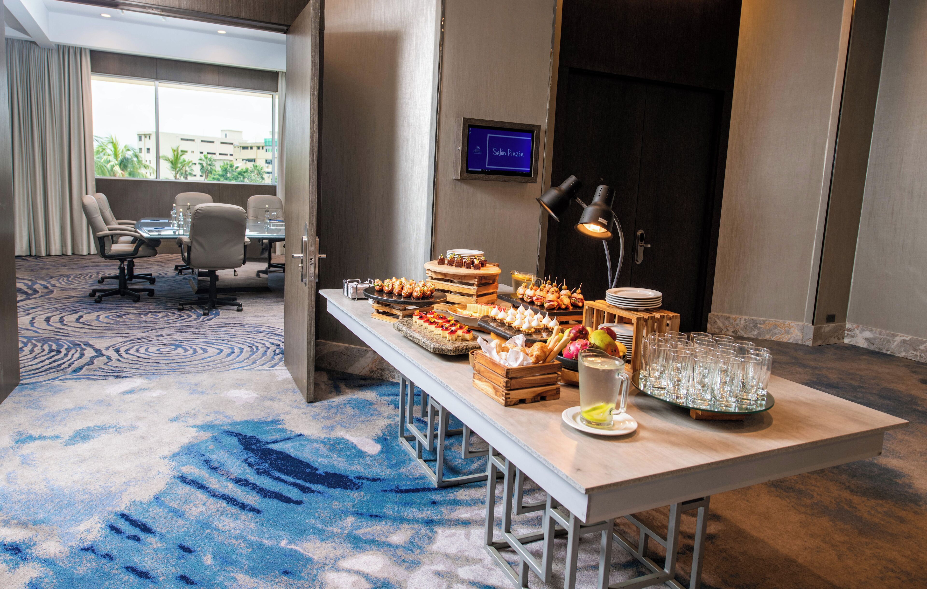 A hotel room with a dining area featuring a buffet table with various snacks and drinks, a television screen, and a view of the city through large windows.