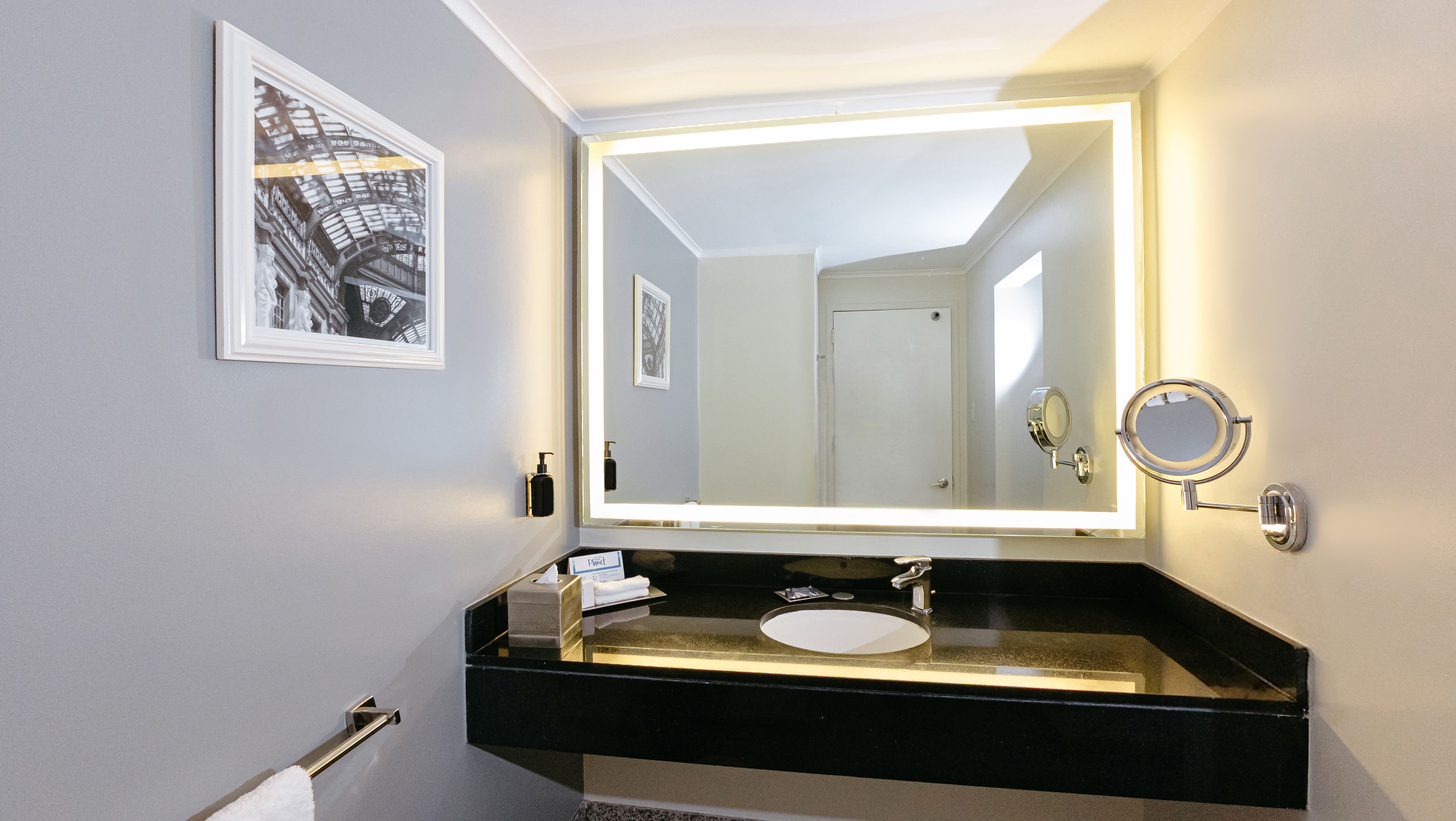 Un baño con tocador negro, un espejo grande con iluminación integrada, un espejo redondo pequeño y un cuadro en la pared.