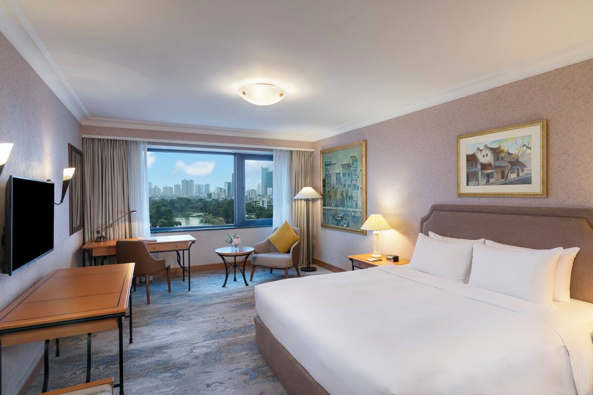 A hotel room with a large bed, a desk, a chair, a small table, a lamp, a painting on the wall, and a view of the city through the window.