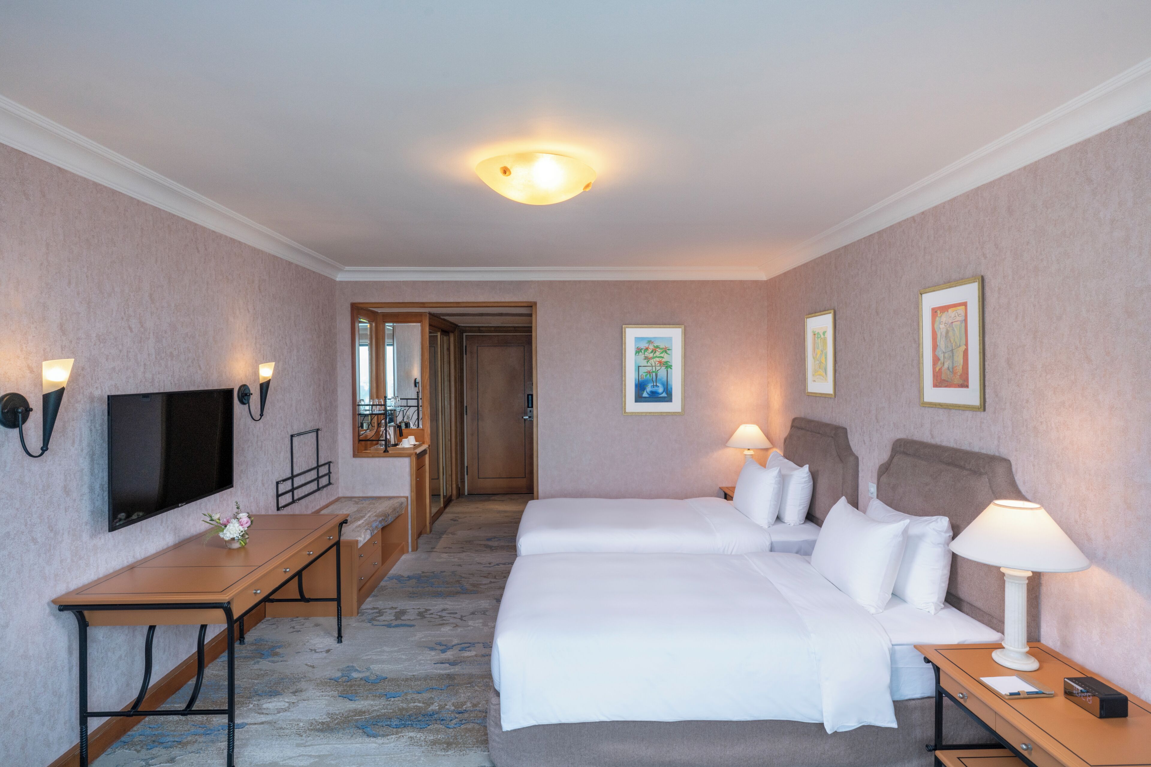 A hotel room with a bed, bedside tables, a desk, a TV, and artwork on the walls.