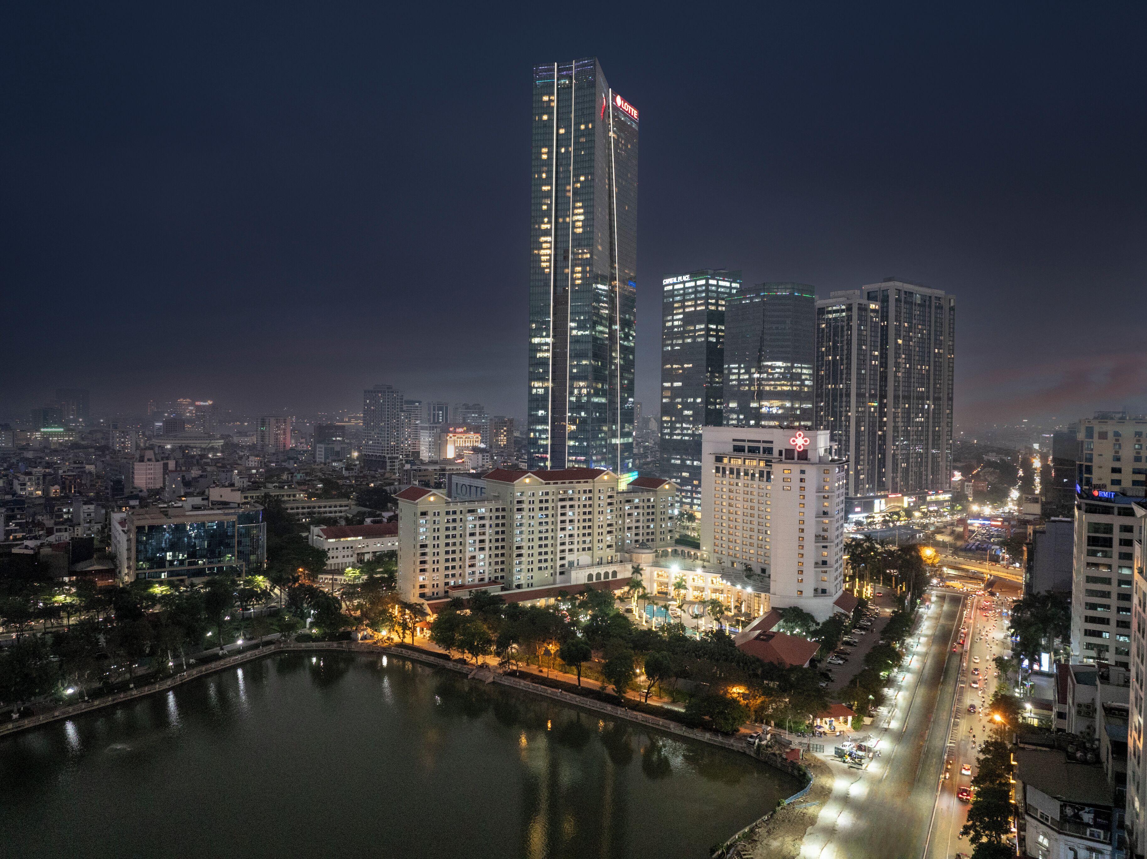 A cityscape at night with illuminated high-rise buildings, a body of water, and a busy road.