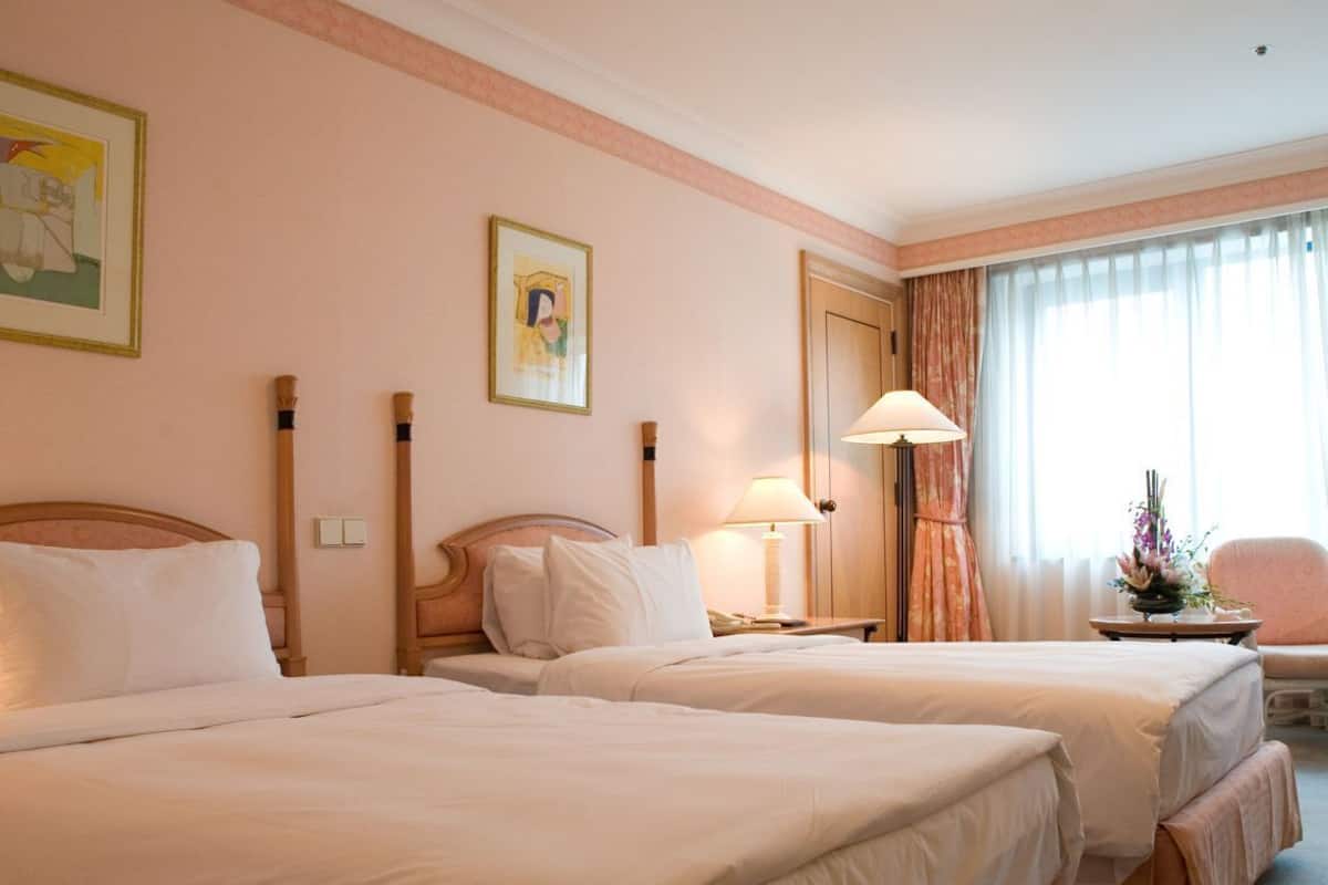 A hotel room with two beds, a bedside table, a lamp, a chair, and a window with curtains.