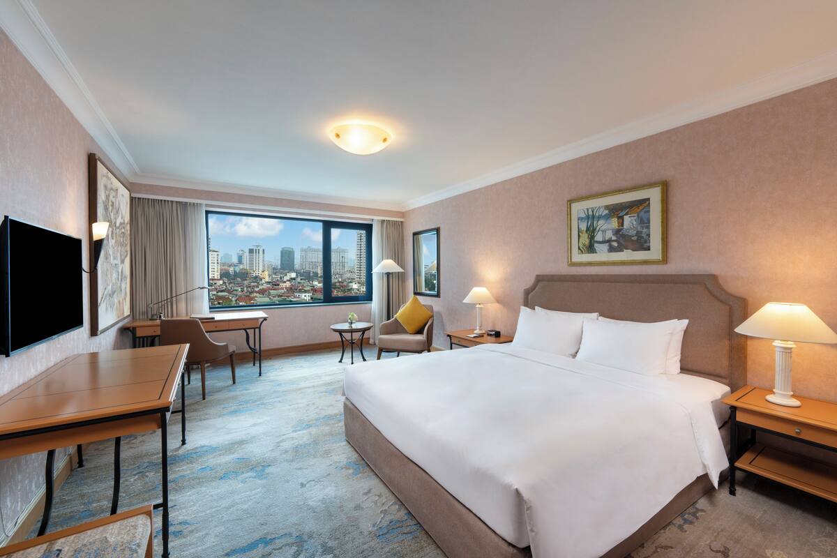 A hotel room with a large bed, a desk, a chair, a TV, and a city view.