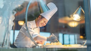 Un chef con uniforme blanco preparando comida en una barra, con una cocina moderna de estilo industrial visible a través de un ventanal.