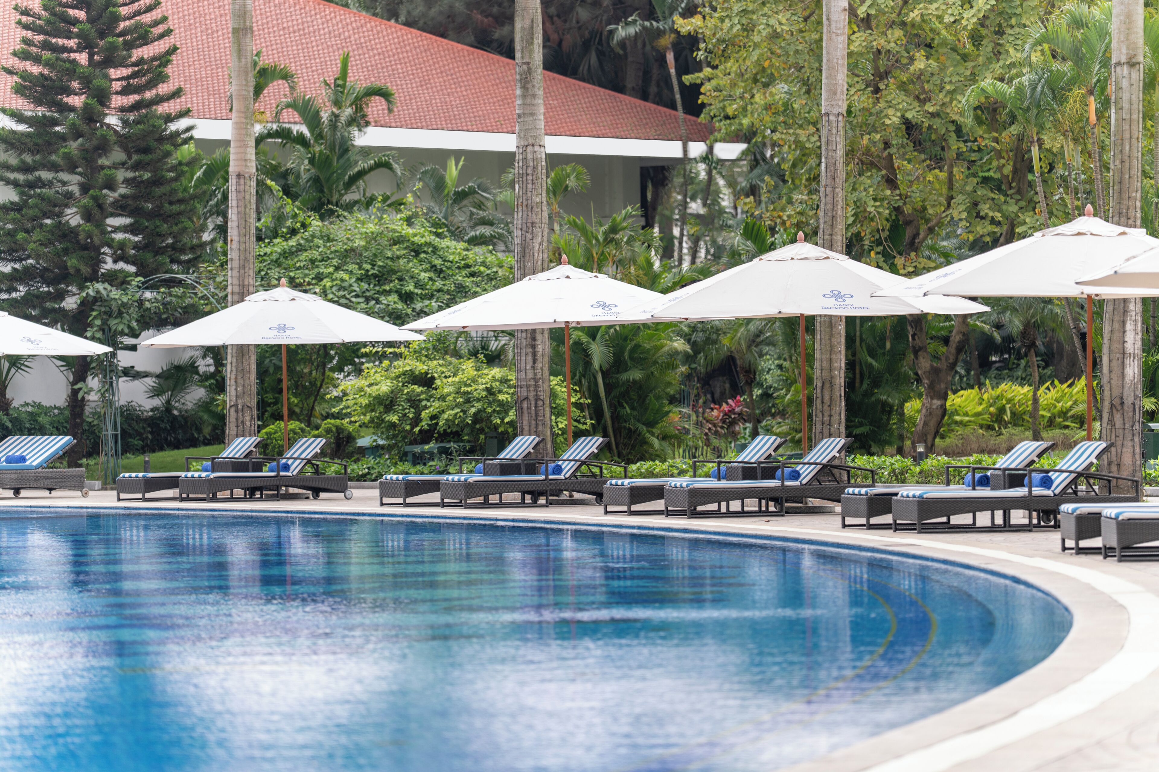 A swimming pool with lounge chairs and umbrellas, surrounded by greenery and a building with a red roof.