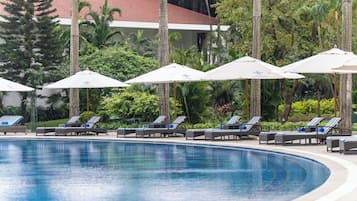 A swimming pool with lounge chairs and umbrellas, surrounded by greenery and a building with a red roof.