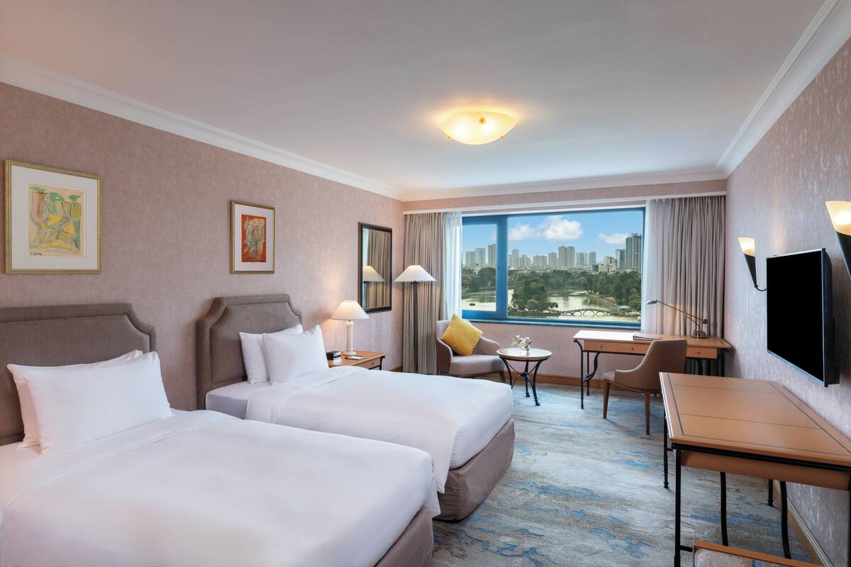 A hotel room with two beds, a desk, a chair, a TV, and a large window with a city view.