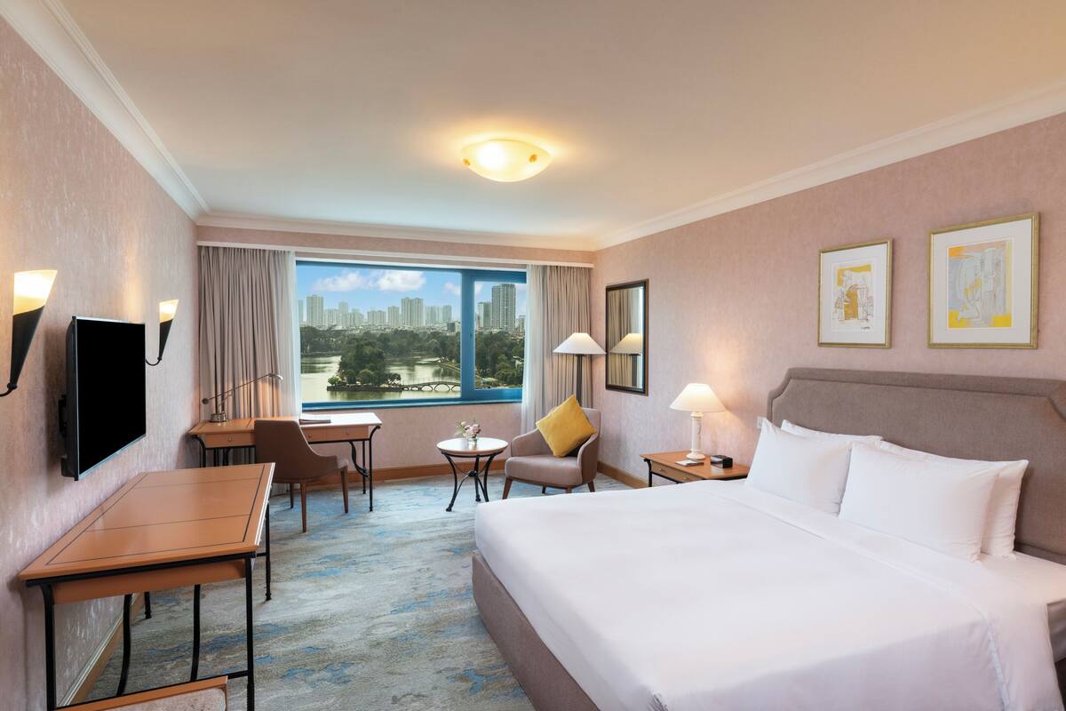 A hotel room with a bed, desk, chair, lamp, and a large window with a city view.