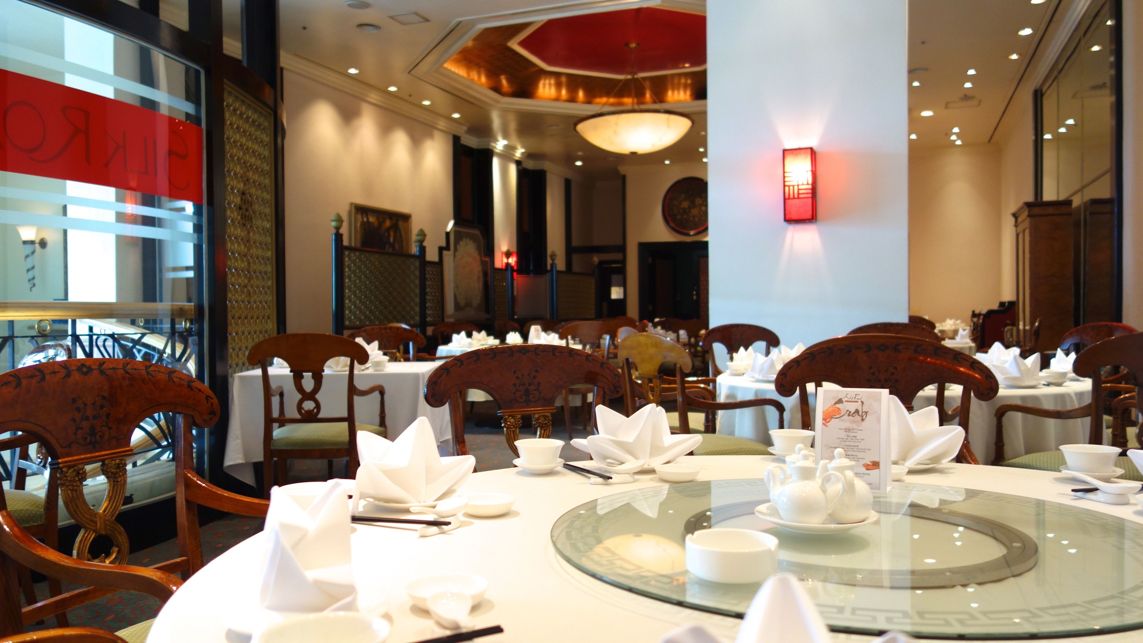 A restaurant with round tables set for dining, wooden chairs, and a glass-top table centerpiece.
