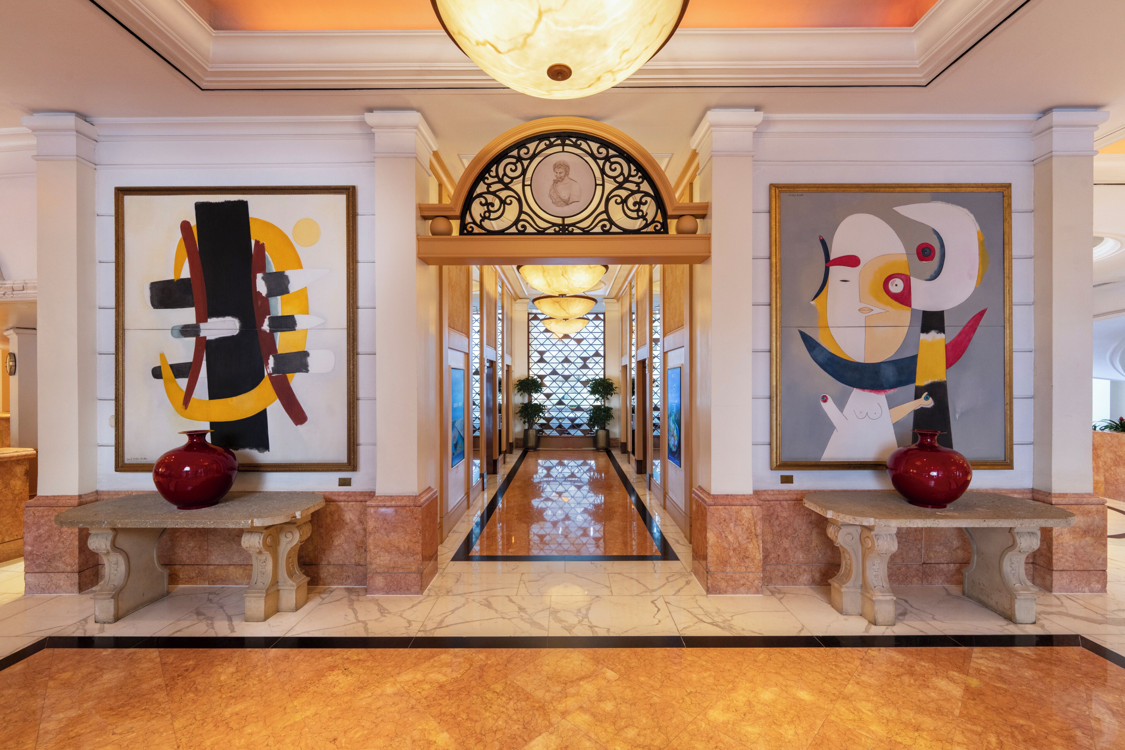 A hallway with marble flooring, two large abstract paintings, a decorative archway, and a chandelier.