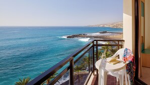 A balcony with a view of the ocean, a white table with a book and glass, and a chair with a colorful blanket. - Sol Tenerife (Arona)