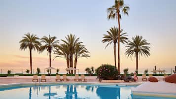 Zona de piscina con sillas de descanso y sombrillas, rodeada de palmeras y una valla.