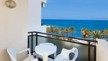 Un balcon avec une table et des chaises blanches, donnant sur une plage bordée de palmiers et sur l’océan.