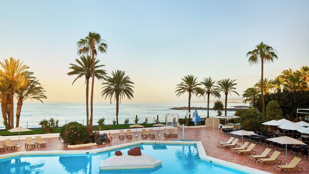 A pool area with lounge chairs and umbrellas, overlooking a beach with palm trees and the ocean. - Sol Tenerife (Arona)