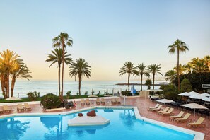 Zona de piscina con sillas reclinables y sombrillas, con vista a una playa con palmeras y el océano.