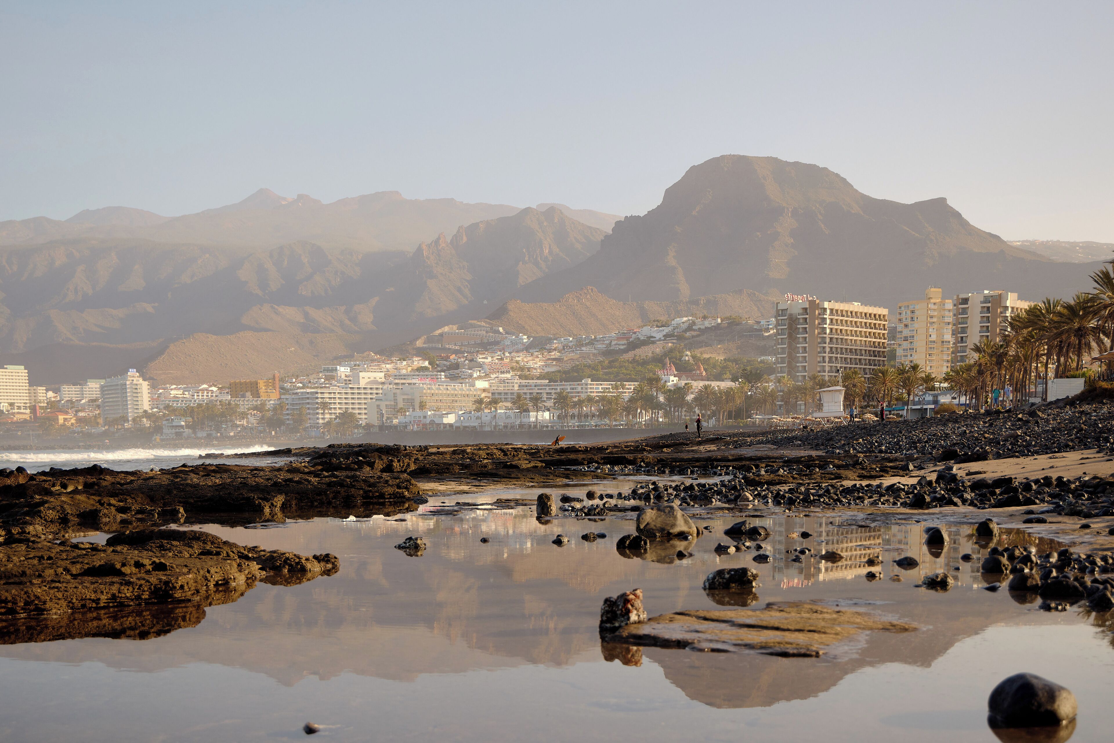 A coastal cityscape with mountains in the background, a beach with rocks, and buildings along the shore.