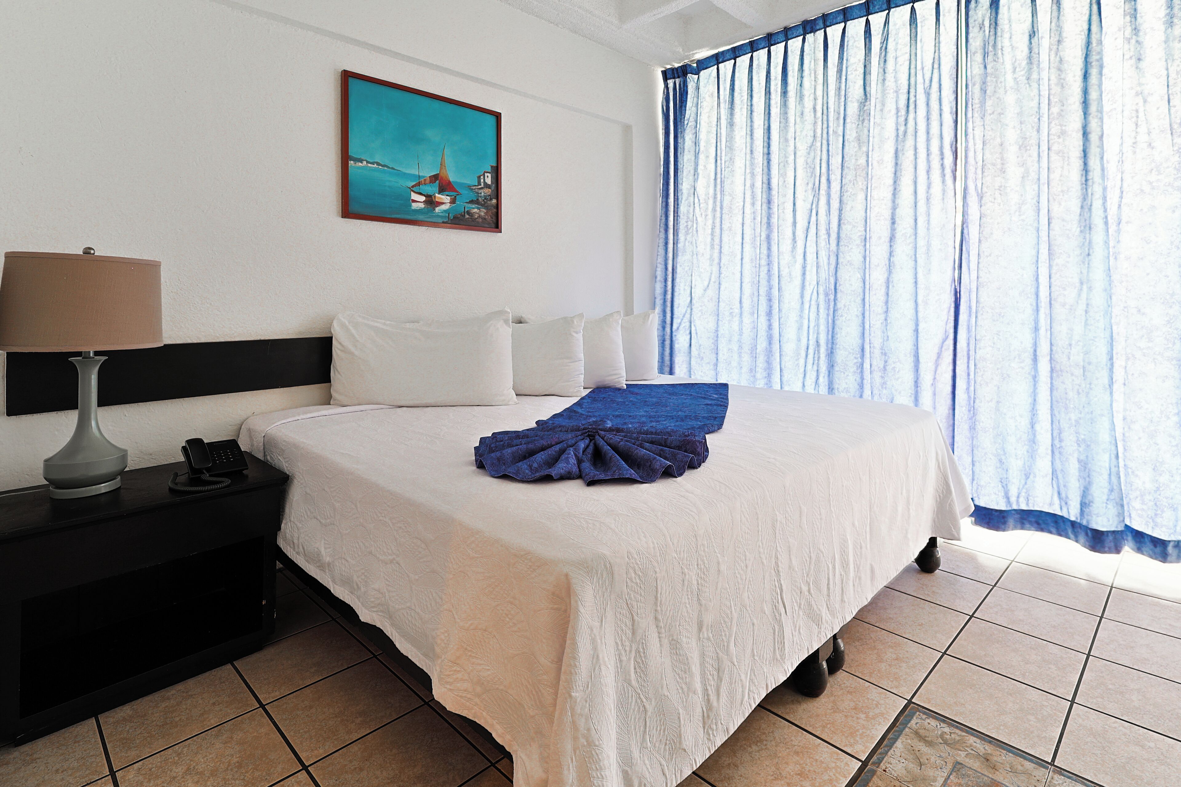 A hotel room with a bed, a nightstand with a lamp, a telephone, and a painting on the wall.