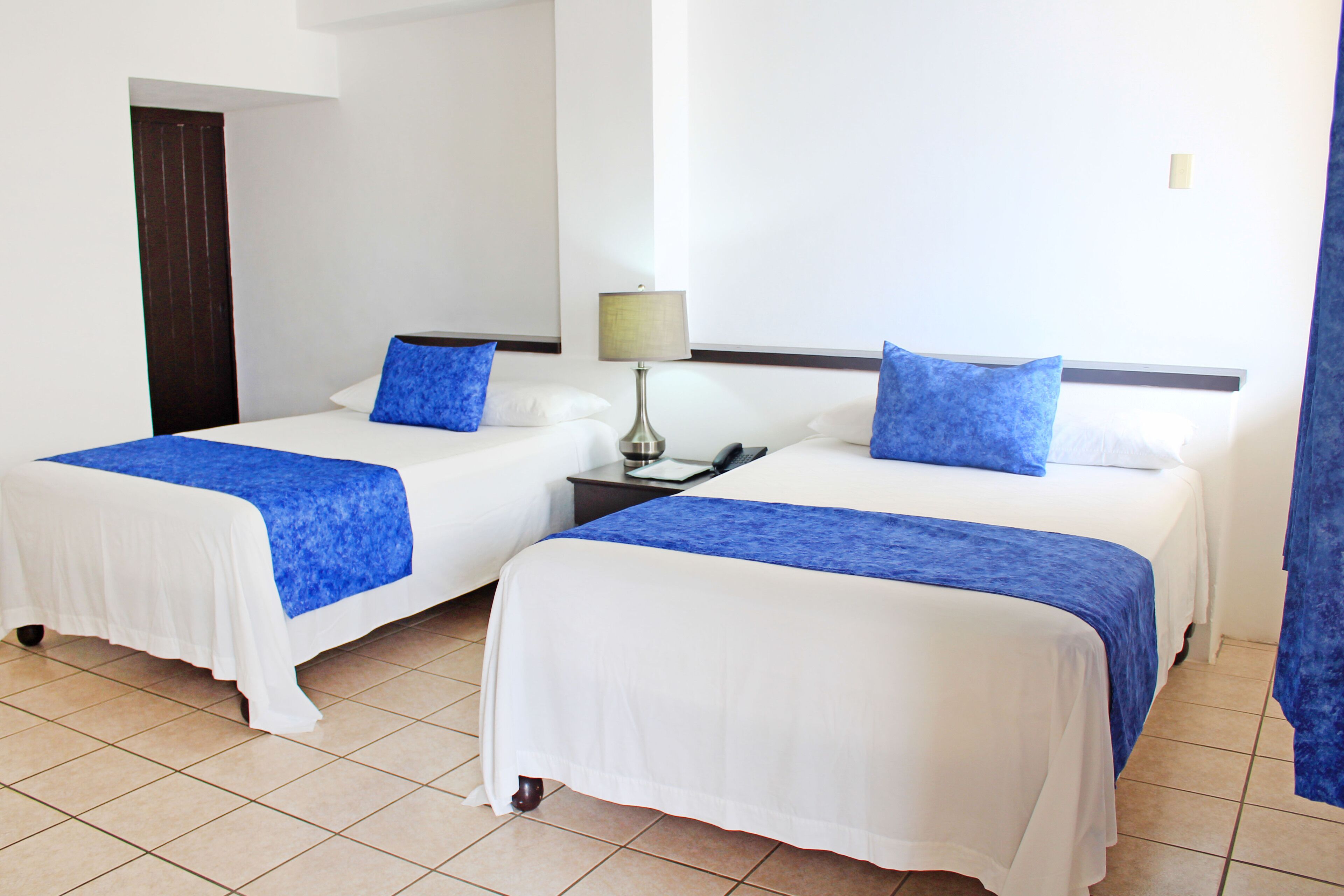 Habitación de hotel con dos camas, cada una con cubrecama blanca y detalles azules, una mesita de noche con lámpara, y una puerta al fondo.