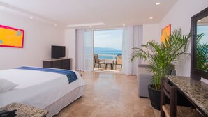 A hotel room with a bed, a TV, a balcony with a view of the sea, and a potted plant.