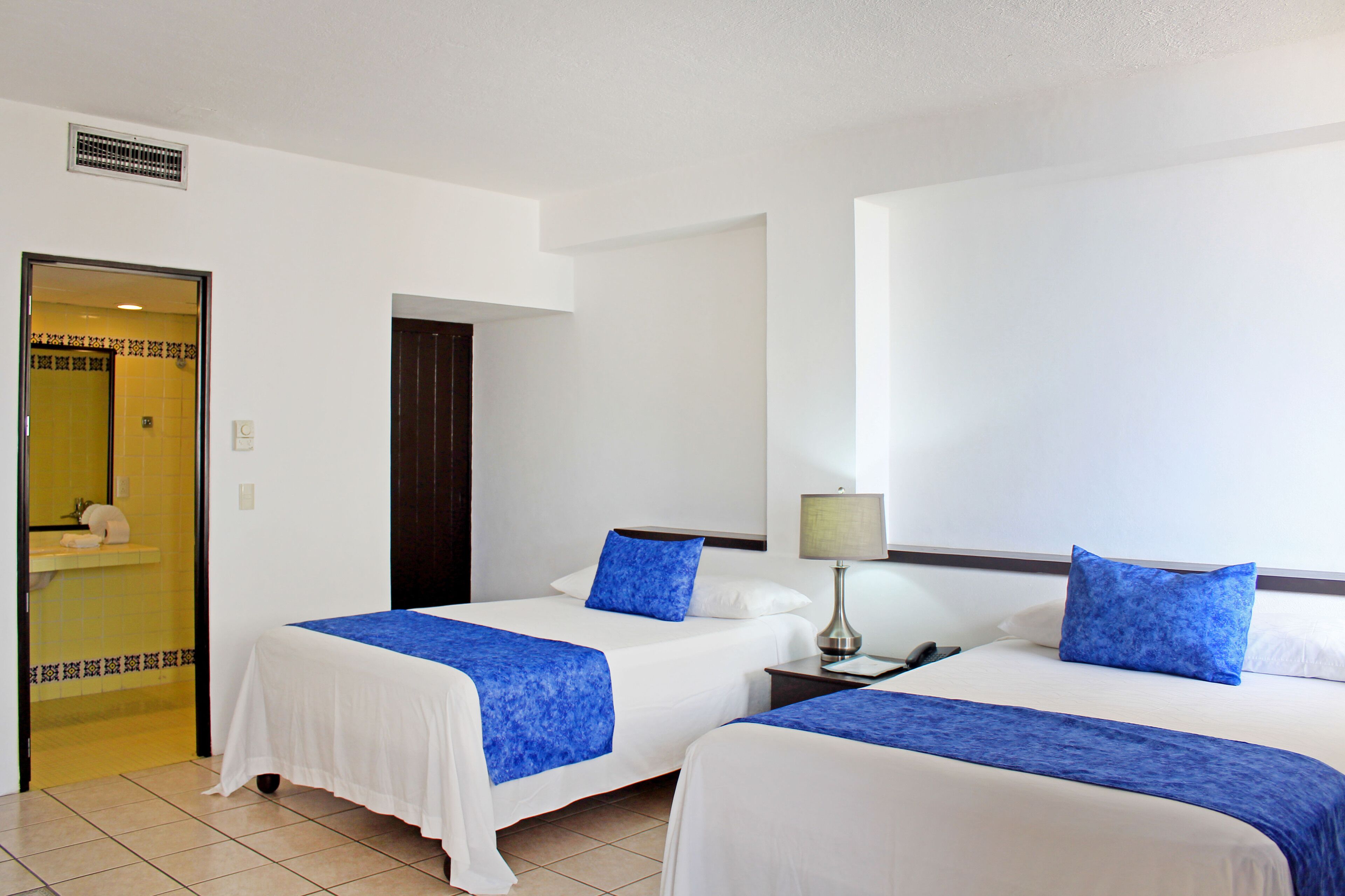 A hotel room with two beds, each with a blue bedspread and white linens. There is a nightstand with a lamp between the beds.