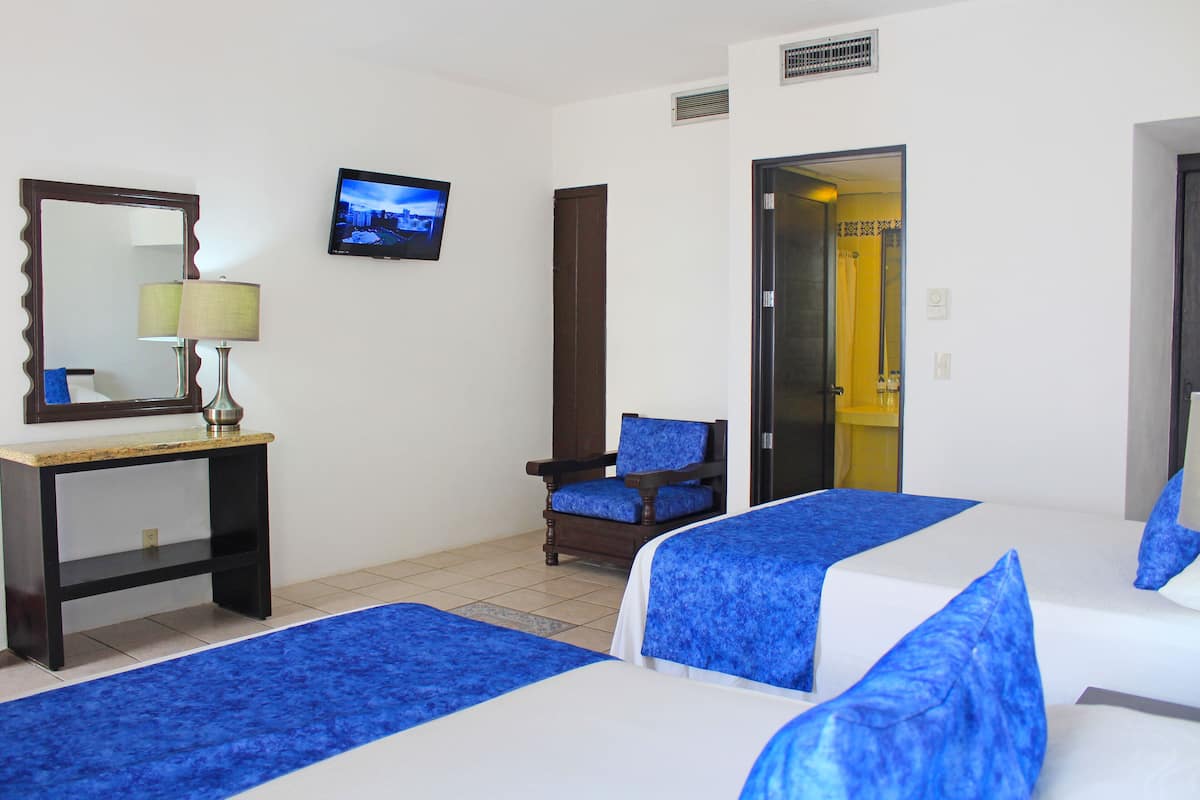 A hotel room with two beds, a desk with a lamp, a chair, a TV, and a door leading to another room.