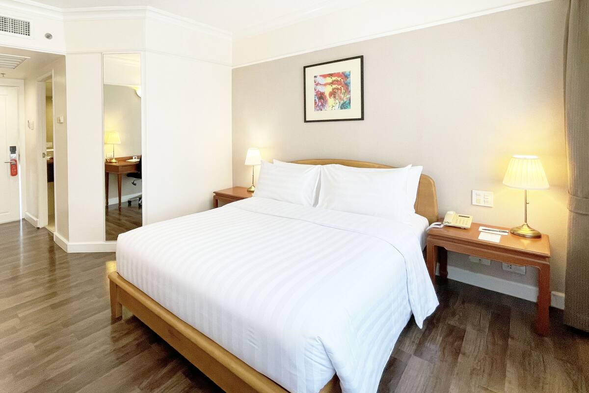 A hotel room with a large bed, wooden headboard, two bedside tables with lamps, a small desk, and a framed picture on the wall.