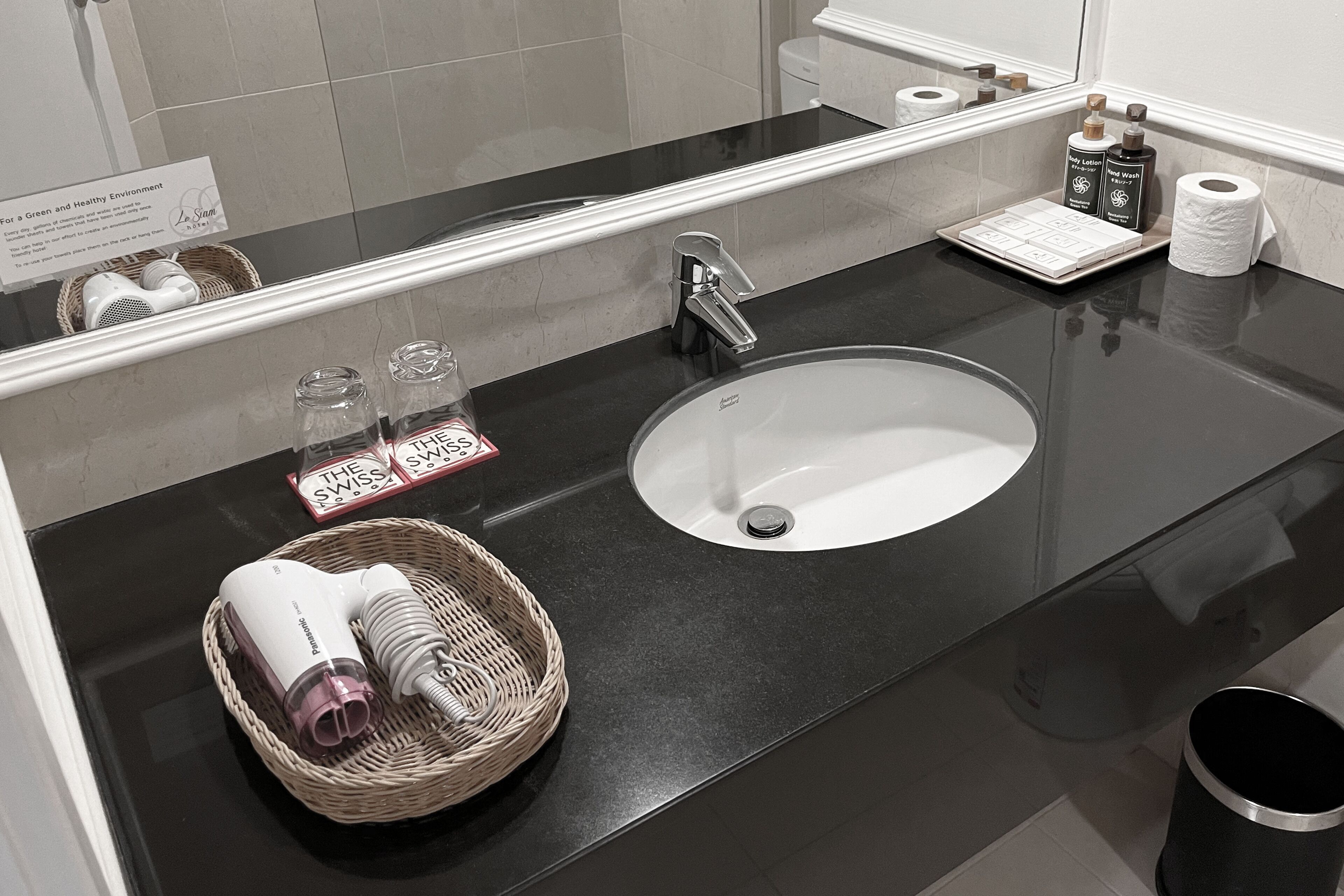 A modern bathroom with a black countertop, a white sink, and a mirror reflecting the room. There are toiletries and a basket with a hairdryer on the counter.
