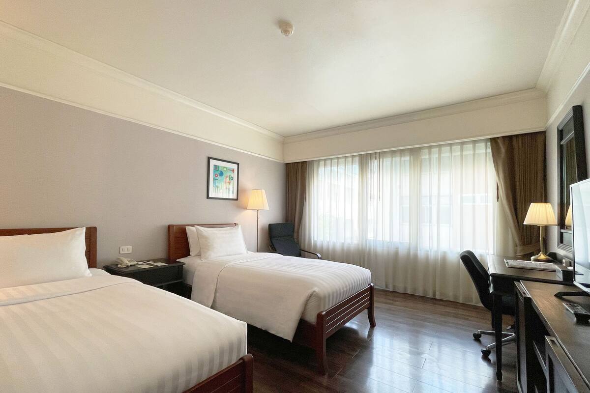 A hotel room with two beds, a desk, a chair, a lamp, and a large window with curtains.
