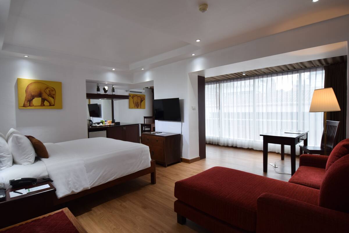 A hotel room with a bed, a desk, a TV, a red sofa, and a large window with curtains.
