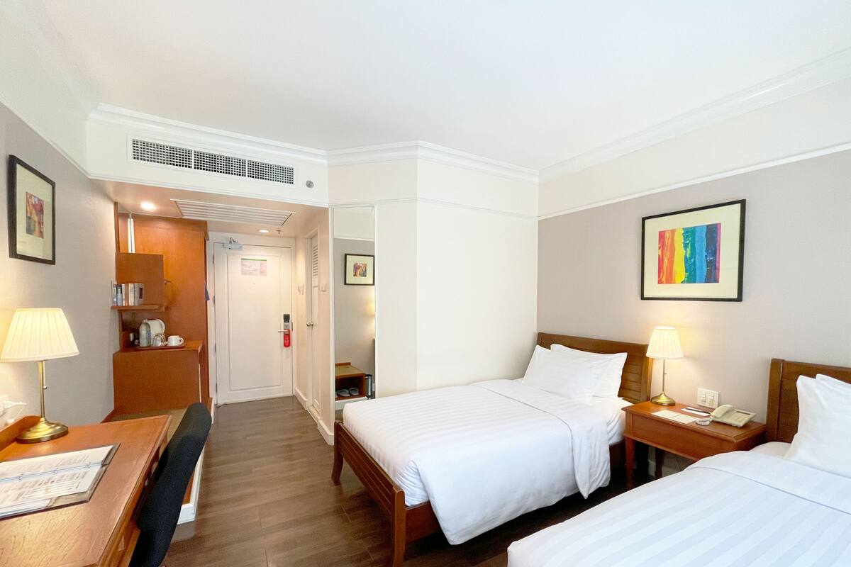 A hotel room with two beds, a desk with a lamp, a chair, a wardrobe, and a door leading to a bathroom.