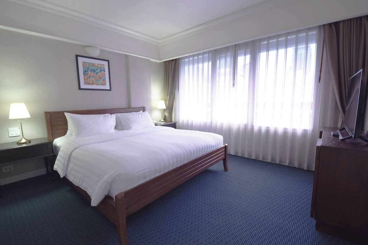 A hotel room with a large bed, two bedside tables with lamps, a wooden headboard, a framed picture on the wall, and a window with curtains.
