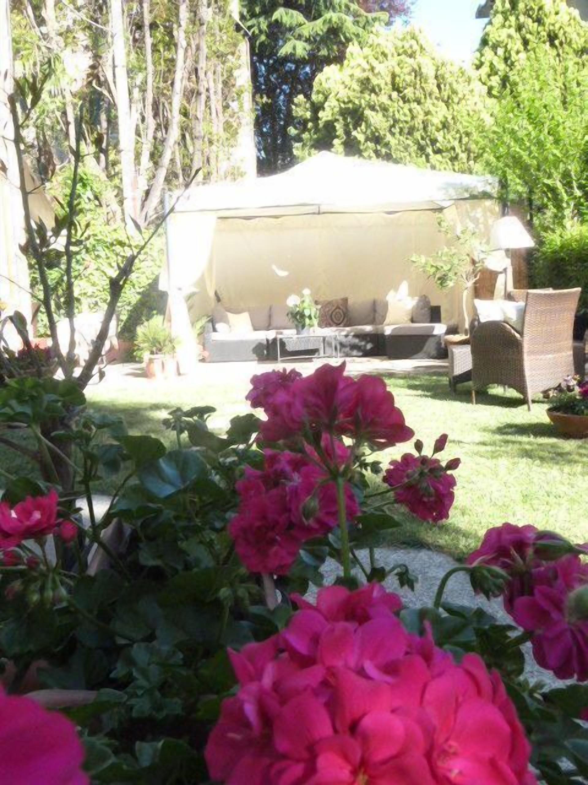 A covered outdoor seating area with a sofa and chairs, surrounded by lush greenery and flowering plants.