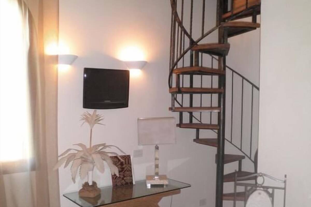 A room with a spiral staircase, a flat-screen TV mounted on the wall, a small table with a plant, and a chair.
