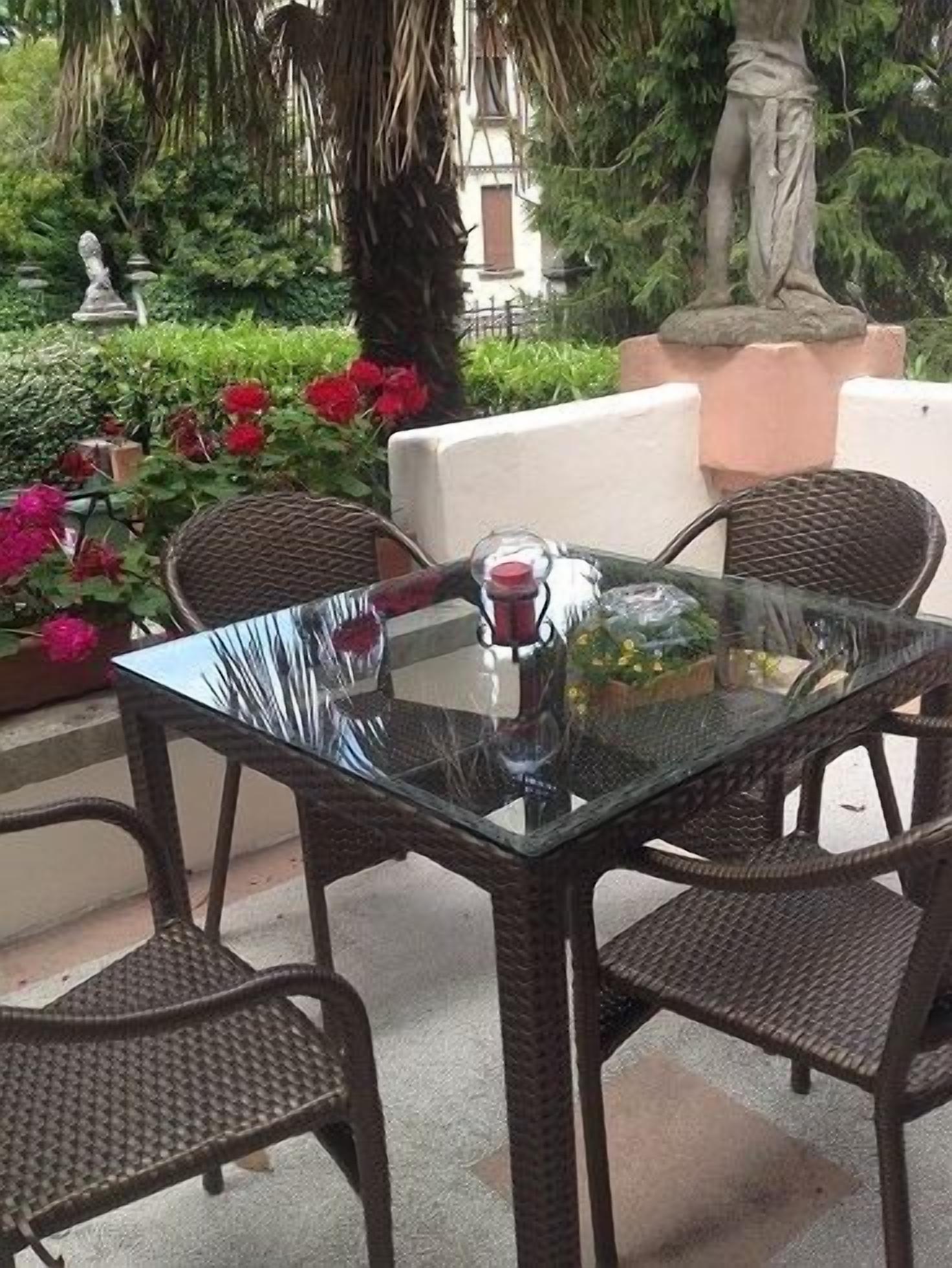 Ein Tisch mit Glasplatte und Korbstühle draußen, im Hintergrund Pflanzen und eine Statue.