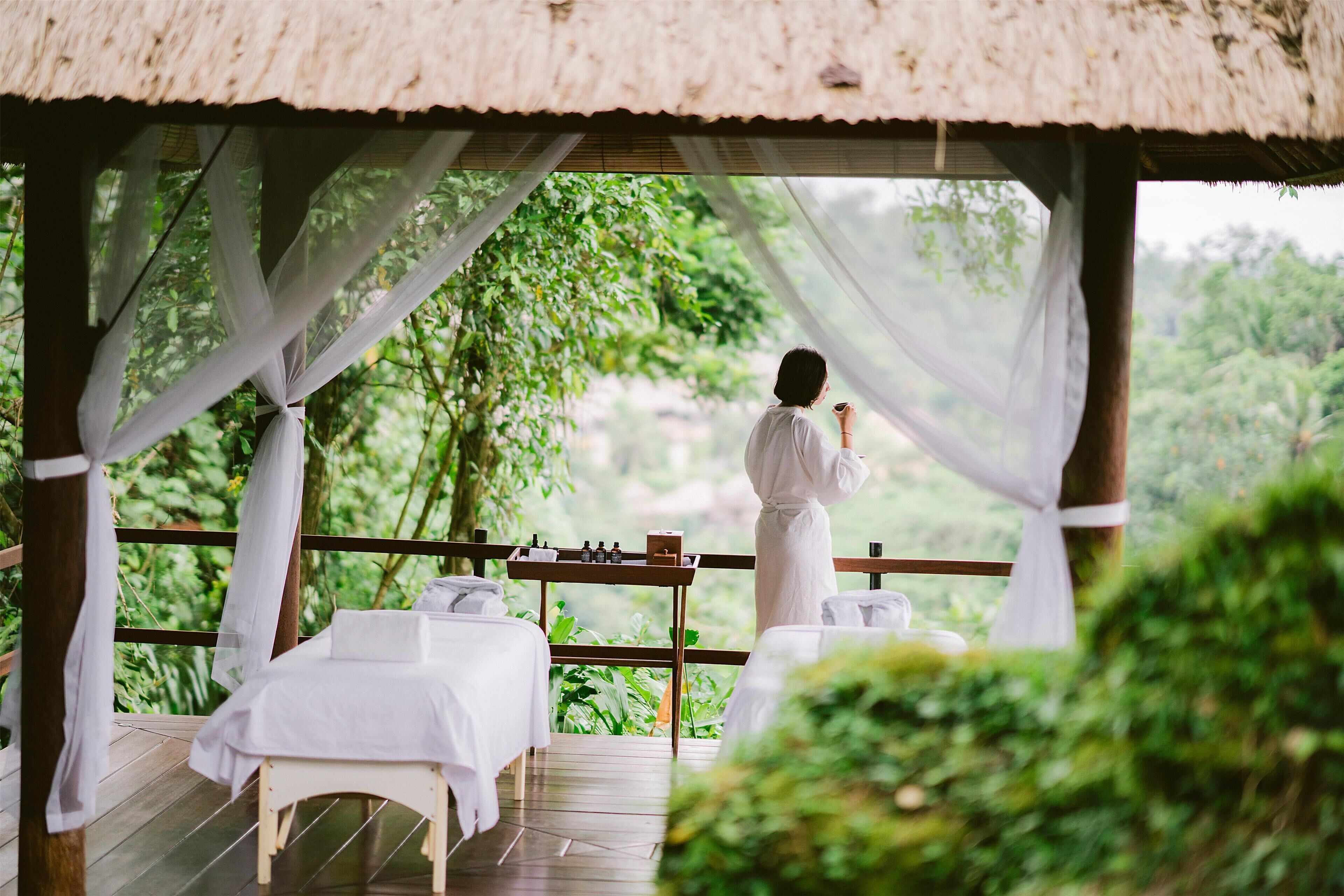 A thatched-roof outdoor seating area with white drapes, wooden tables, and benches, overlooking a lush green landscape.