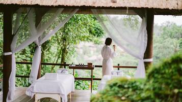A thatched-roof outdoor seating area with white drapes, wooden tables, and benches, overlooking a lush green landscape.