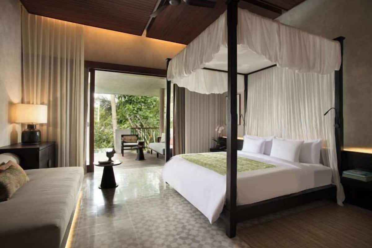 A four-poster bed with white linens, a bedside table with a lamp, and a view of greenery outside.