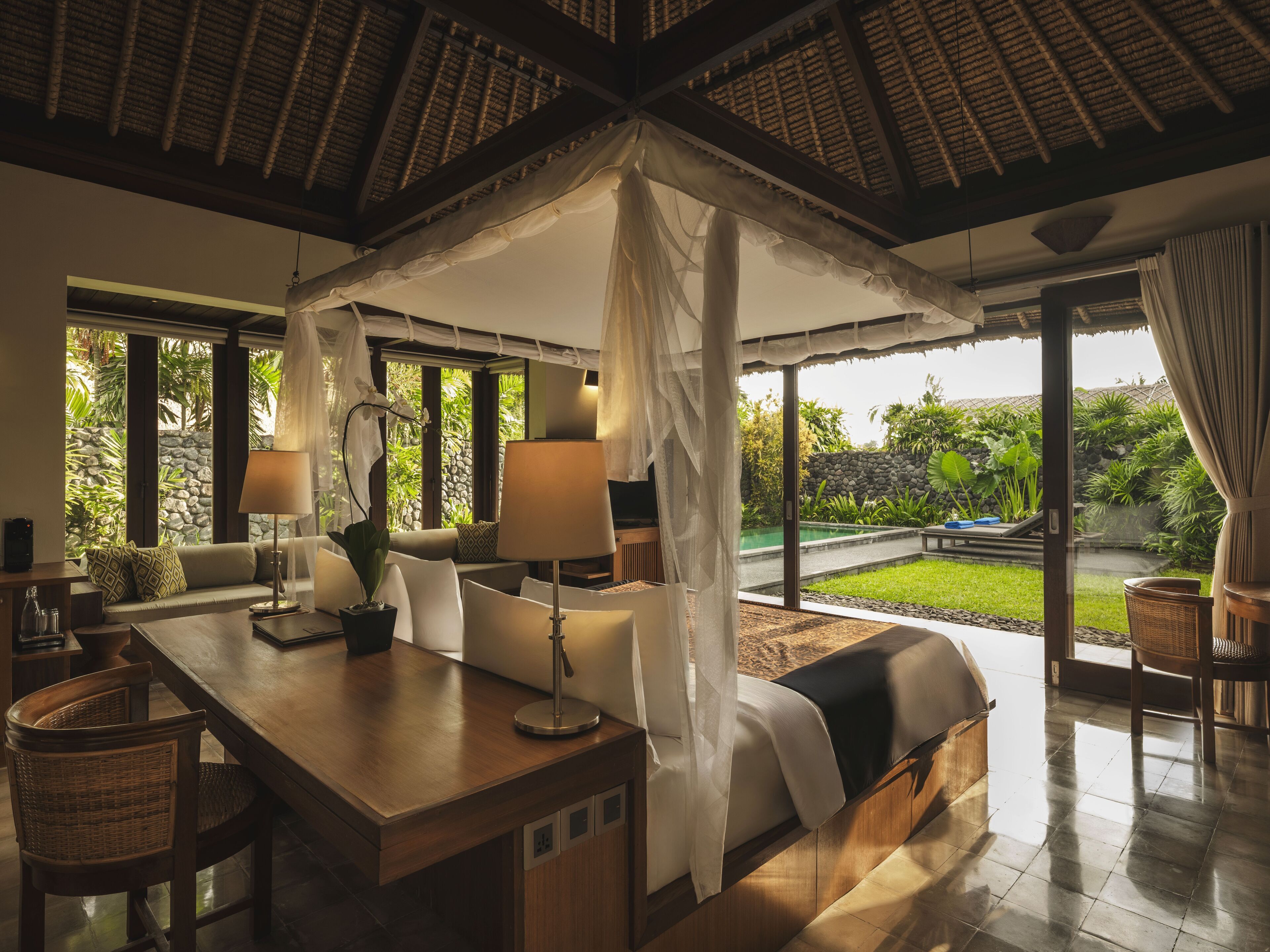 A room with a bed, a dining table, and chairs, a large window with a view of a garden and pool.