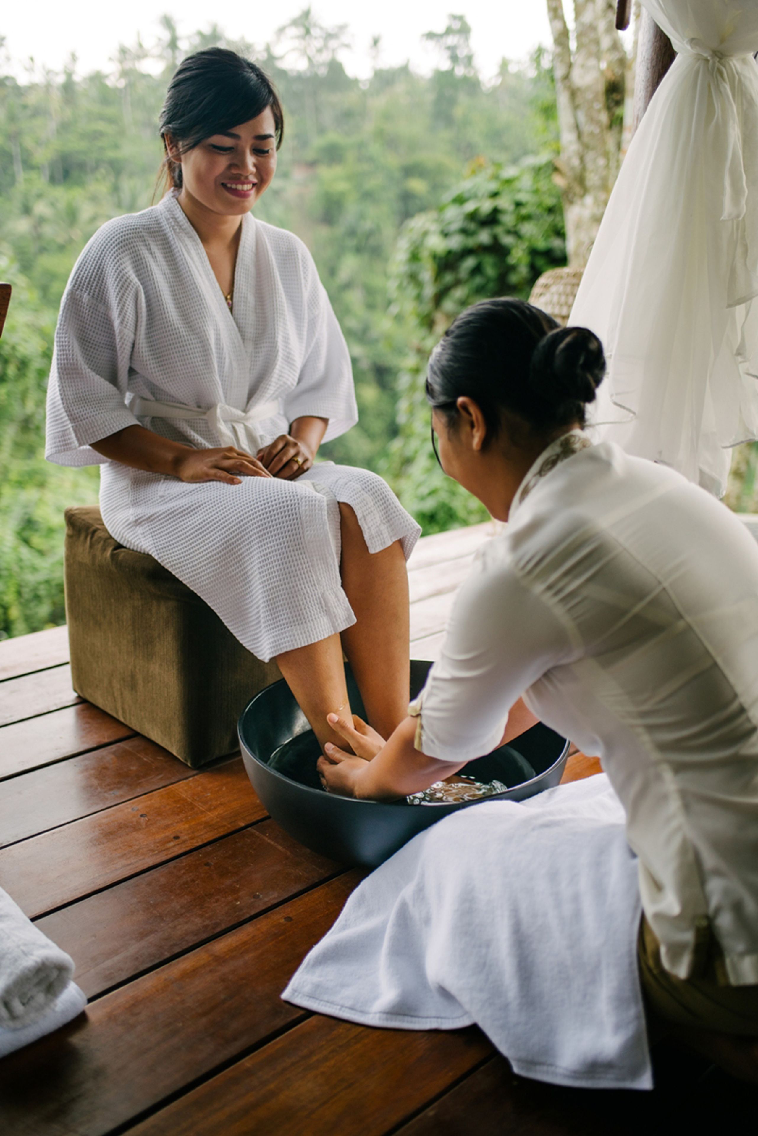 A person receiving a foot soak in a wooden basin outdoors with a lush green background.