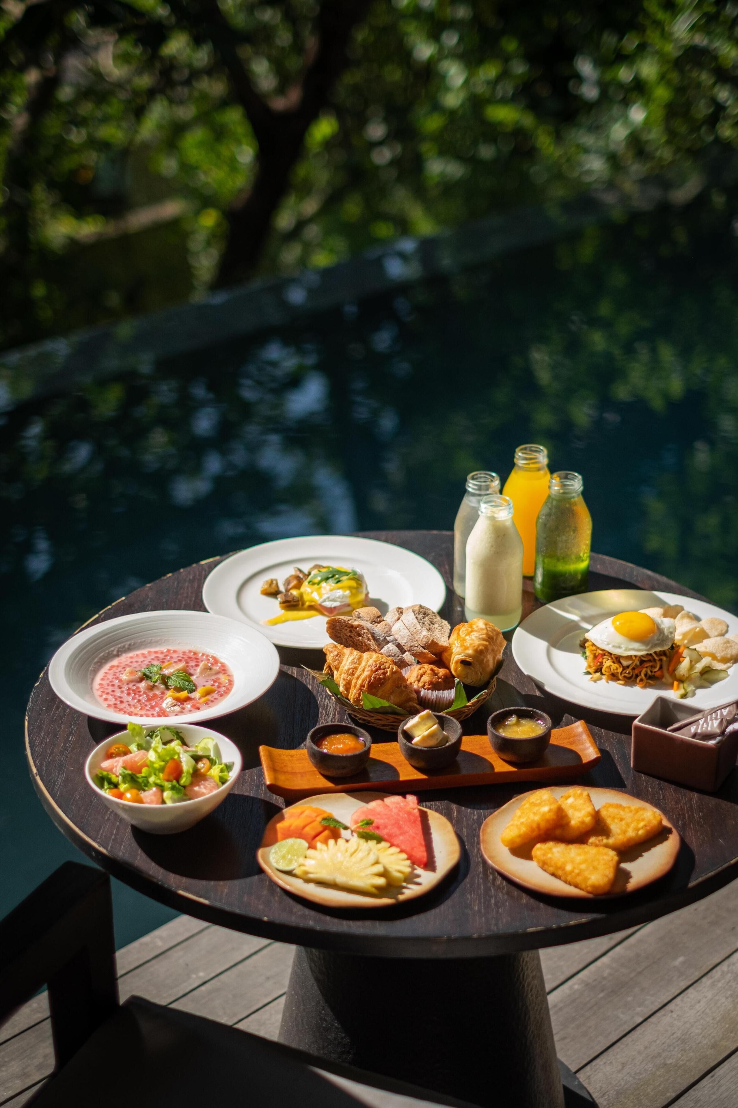 A dining table with various dishes and drinks, set against a backdrop of a body of water and greenery.