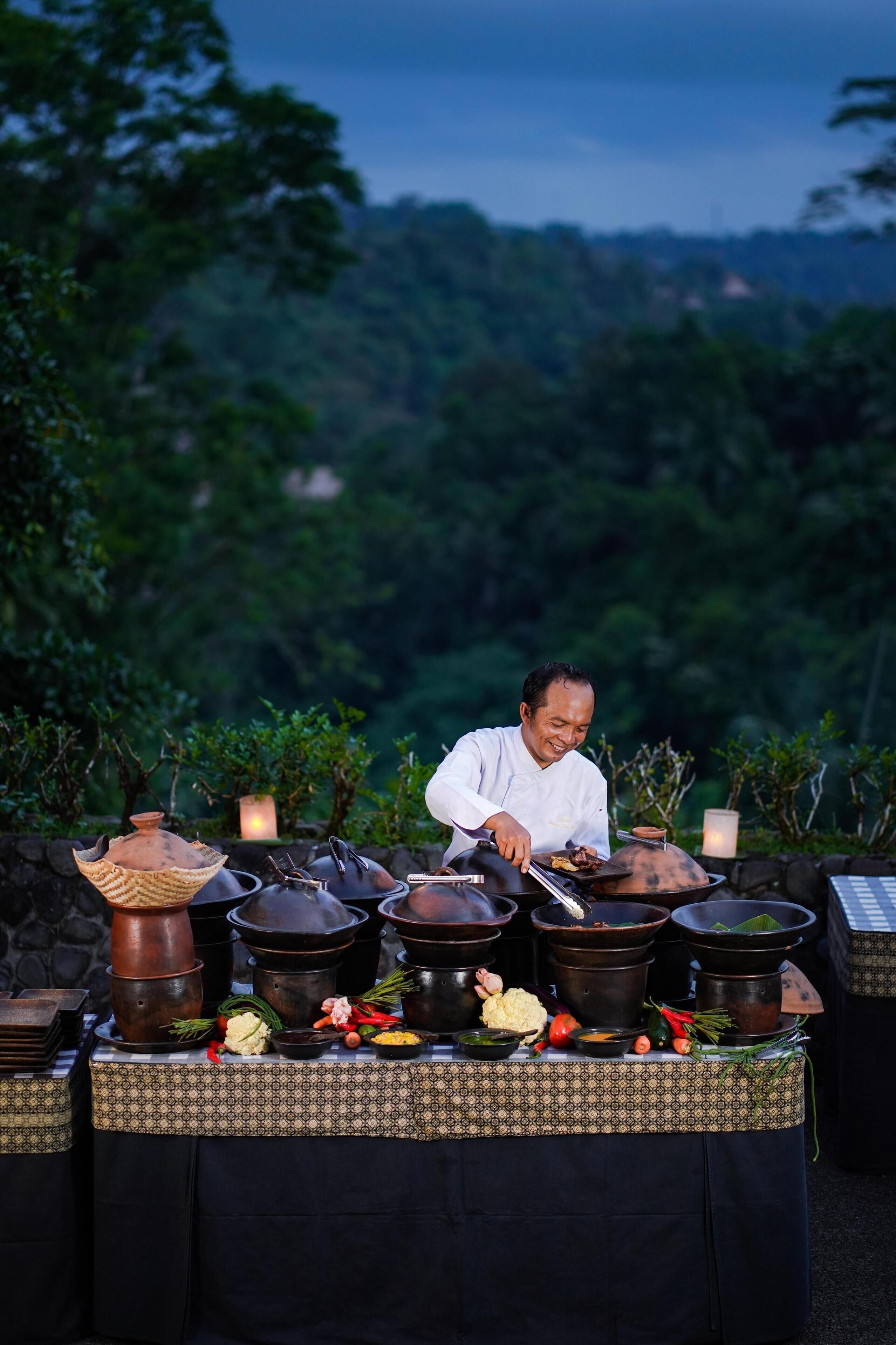 A chef preparing food outdoors with multiple cooking pots and fresh ingredients on a table.